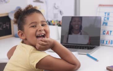 6 years old girl studying at home online, using a laptop computer. Elementary student and her teacher using a video chat app to communicate remotely. The happy mixed-race girl with hand on chin looking over the shoulder at the camera with a laughing face. Her private teacher showing a notebook on the laptop's screen in the background. Distance learning during the Coronavirus outbreak.